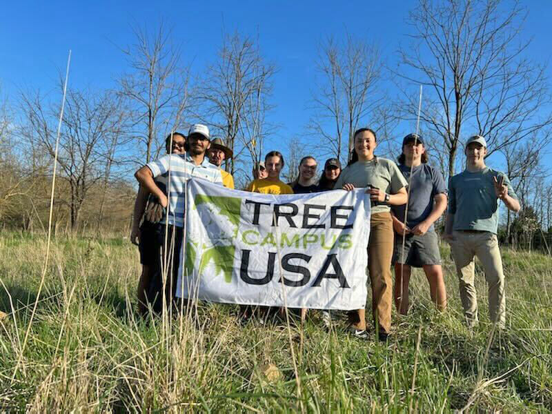 Photo of students standing in field holding tree campus USA sign in front of trees they just planted.