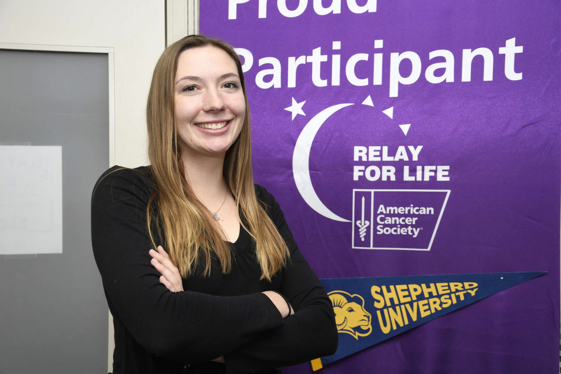 Photo of Aly Nazarok standing in front of Relay For Life banner smiling with her arms crossed.