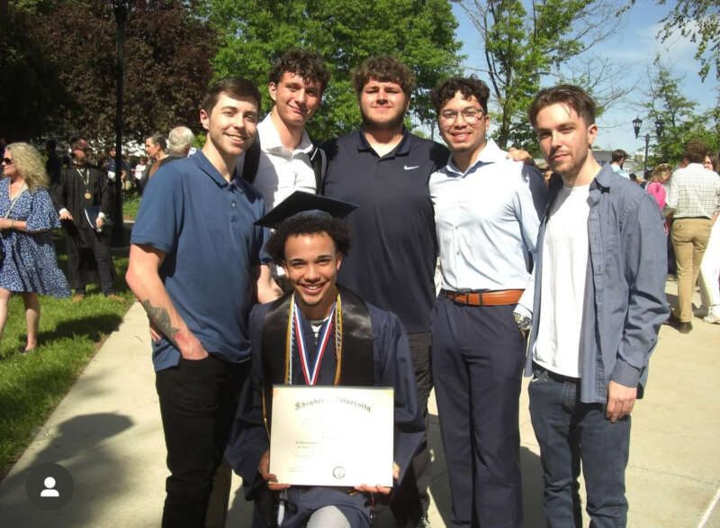 Members of a fraternity dressed up stand behind their fraternity brother dressed in graduation cap and gown holding his Shepherd diploma