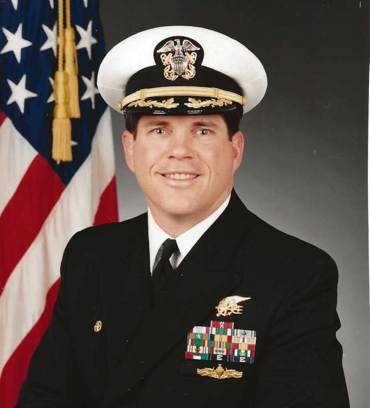 Commander Tom Deitz US Navy Ret in full dress uniform with medals and insignia standing in front of American flag