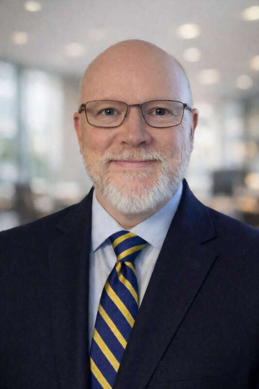 Person wearing a navy suit jacket, light blue dress shirt, and blue and yellow striped tie standing in an office setting with soft background lighting.